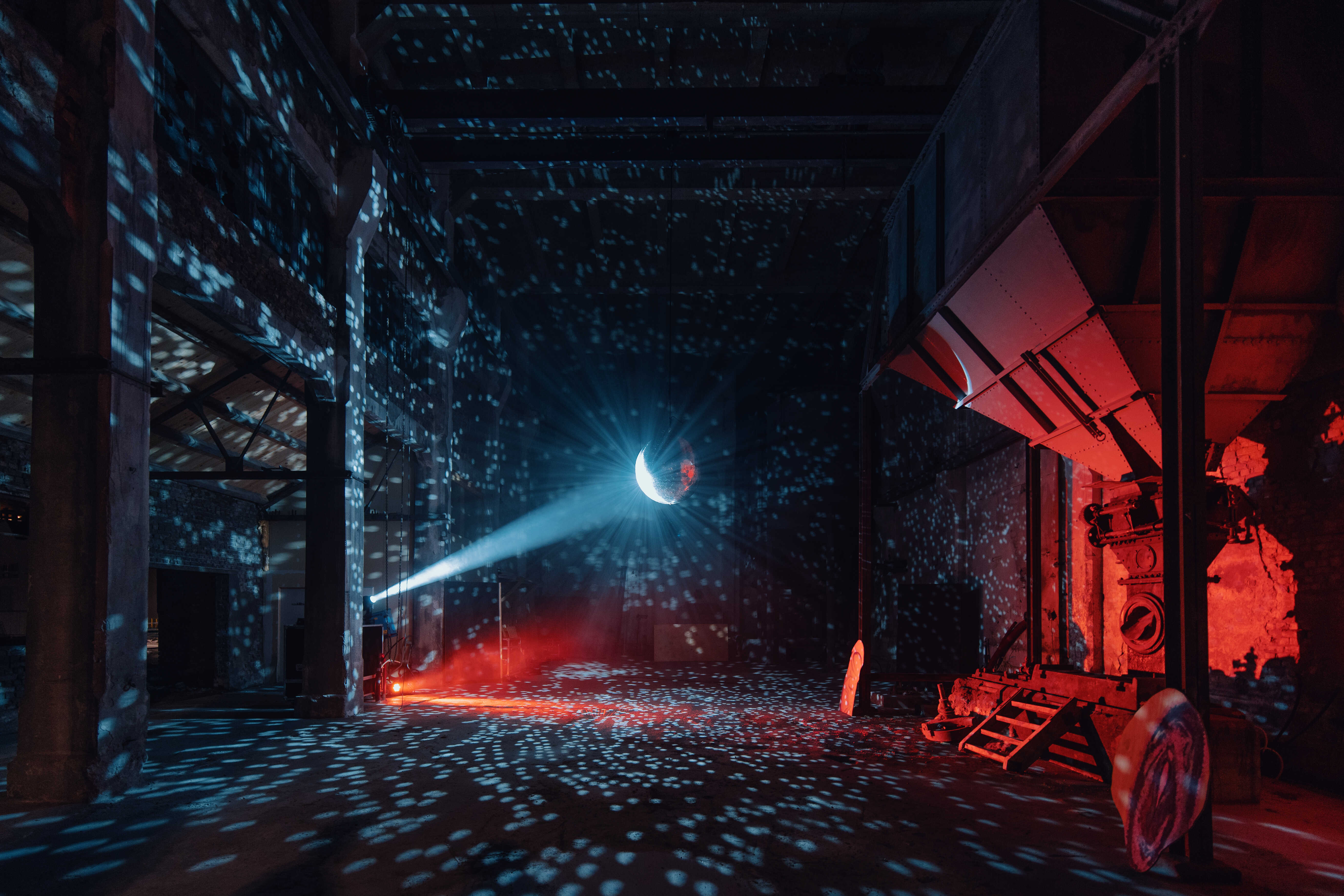 En stor diskokule opplyst av en lyskaster henger i et mørkt industrilokale. Foto.