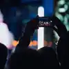 Nært detaljbilde av en person som løfter mobilen sin høyt opp for å ta et bilde. Motlys fra scenen gjør at man kun ser silhuett av fotografen og andre publikummere rundt. Foto.
