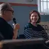 Nærbilde av forfatter Frode Grytten og ordstyrer Elise Winterthun som sitter på scenen på Sentralbadet litteraturhus. Frode snakker i en mikrofon og Elise smiler mot ham. I en vinduskarm i bakgrunnen står en kaffekjele med store vinger. Foto.