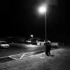 Det er natt, og et par står tett omslynget under en lyktestolpe på en gressplen ved en tom parkeringsplass. Foto.