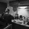 En kvinne står med ryggen til kamera ved kjøkkendisken i byssen på Epos. På disken står kaffetrakter, kaffekanner og vannkoker, og over disken er et vindu som viser Sørfjorden. Foto.